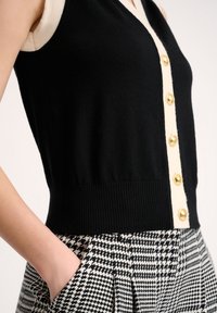 Černý pletený top bez rukávů s kontrastním krémovým zapínáním na knoflíky a zlatými knoflíky, spárovaný s kalhotami v houndstooth vzoru.