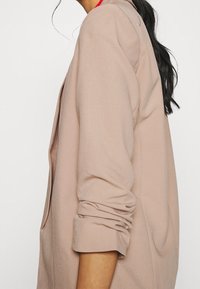 Blazer beige avec des manches retroussées, fabriqué en tissu doux. Présente une coupe décontractée et un design minimaliste. Aucun matériel ou motif visible.