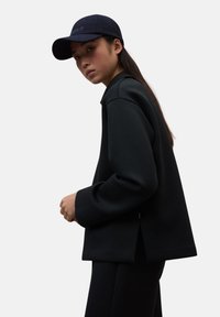 Joven mujer con cabello largo y oscuro que lleva una gorra negra, una chaqueta negra de gran tamaño y pantalones a juego, mirando de lado contra un fondo simple.