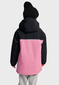 Veste imperméable noire et rose avec capuche ; elle possède des poignets élastiques, une bande réfléchissante et une texture lisse. Bonnet tricoté noir porté.