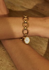 Montre en métal doré avec un boîtier circulaire, un cadran blanc et des index horaires. Le bracelet est composé de segments ronds et ovales entrelacés.