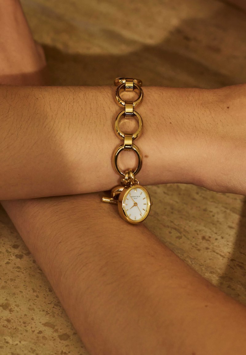 Montre en métal doré avec un boîtier circulaire, un cadran blanc et des index horaires. Le bracelet est composé de segments ronds et ovales entrelacés.