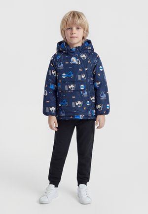 Garçon blond debout portant une veste à capuche bleu marine avec des imprimés de véhicules de chantier, un pantalon noir et des baskets blanches sur fond blanc.