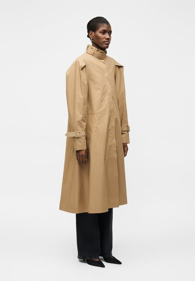 Femme portant un long trench-coat beige avec des brides à boucle aux manches, un pantalon noir à jambes larges et des chaussures noires pointues, debout sur fond blanc.