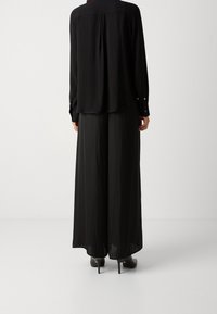 Blusa nera a maniche lunghe con schiena plissettata, abbinata a pantaloni neri a gamba larga. Texture liscia; dettagli lucidi sui bottoni. Scarpe con tacco.