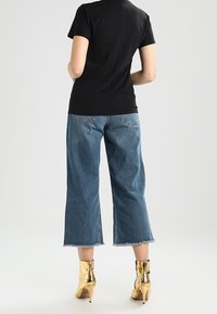 Černé tričko s krátkým rukávem, modré džíny s vysokým pasem a zkrácenou délkou s třepením na lemu, a lesklé zlaté kotníkové boty. Neutrální pozadí.