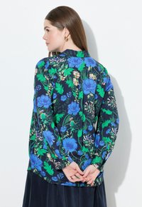 Donna con lunghi capelli castani che indossa orecchini a cerchio dorati e una blusa blu navy con vivaci motivi floreali blu e verdi, con le mani unite dietro la schiena.