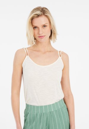 Young woman with short blonde hair wearing a white sleeveless top with double straps and green striped pants, facing forward.