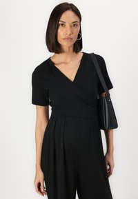 Combinaison noire à manches courtes, en tissu texturé, avec une taille cintrée, associée à un petit sac à main noir.