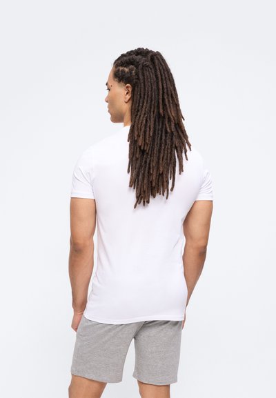 Άνδρας με μακριά dreadlocks φορά λευκό μπλουζάκι και γκρι σορτς, στέκεται γυρισμένος με την πλάτη προς ένα απλό φωτεινό φόντο.