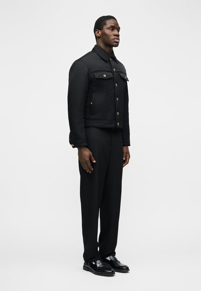 Άνδρας στέκεται φορώντας μαύρο σακάκι με κουμπιά και τσέπες στο στήθος, μαύρο παντελόνι και μαύρα δερμάτινα παπούτσια, σε απλό φωτεινό φόντο.