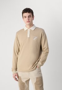 Polo de culoare bej cu mâneci lungi, cu un guler crem, placă cu trei nasturi și logo cu scut pe piept. Material neted și design ajustat.