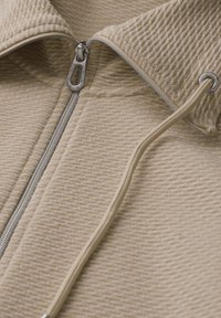 Chaqueta beige texturizada con cremallera y cuello alto, que presenta una cremallera plateada y detalles de cordón en un color a juego.