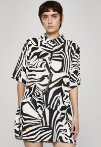 Jeune personne portant une robe chemise à manches courtes avec un motif abstrait noir, blanc et marron, debout devant un fond uni.