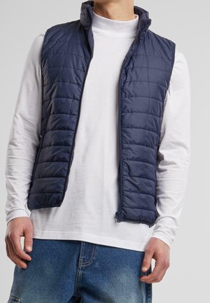 Mann trägt eine dunkelblaue gesteppte ärmellose Weste über einem weißen Langarmshirt und blauen Jeans vor einem einfarbigen grauen Hintergrund.