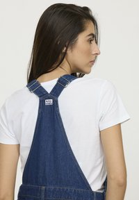 Salopette en denim avec des bretelles bleues réglables et des éléments en métal. Présente une étiquette à l'arrière et un t-shirt blanc uni en dessous. Texture lisse.