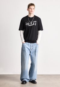 Μαύρο μπλουζάκι με λευκό λογότυπο, τοποθετημένο πάνω από ένα ανοιχτόχρωμο μακρυμάνικο πουκάμισο. Χαλαρές ανοιχτόχρωμες μπλε τζιν παντελόνες, μαύρα παπούτσια. Μινιμαλιστικός σχεδιασμός, άνετη εφαρμογή.