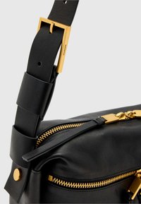 Črna usnjena torba z zlatim kovinskim zadrgo, debelim trakom z zlato zaponko in poudarkom zlatega okovja. Teksturirana površina.