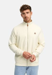 Joven con una chaqueta crema de cremallera y un pequeño parche redondo en el pecho, ajustándose el puño, de pie ante un fondo liso.