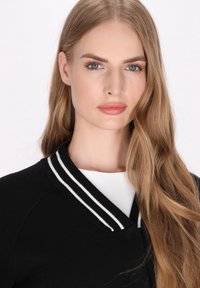 Schwarze Strickjacke mit einem weißen, gestreiften Rippkragen, leichtem Strickstoff und Rundhalsausschnitt. Die Haare sind lang und wellig. Neutraler Hintergrund.