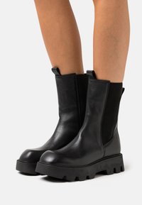 Black leather ankle boots with elastic side panels, round toe, and thick, textured rubber sole featuring deep grooves for traction.
