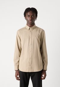 Cămașă butonată bej deschis, cu guler, mâneci lungi și nasturi în față. Prezintă un broderie discretă a logo-ului pe piept și o texturăfină.