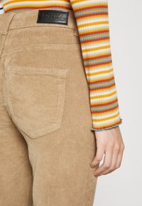 Pantalon en velours côtelé beige avec une surface texturée et une poche arrière. Étiquette en cuir noir sur la taille. Chemise à manches longues rayée colorée visible.