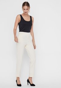 Canotta nera aderente abbinata a pantaloni a sigaretta crema. I pantaloni hanno una texture liscia, con risvolti e una vita piatta. Tacchi neri completano il look.