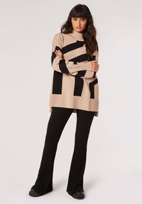 Suéter de punto beige con rayas negras gruesas, cuello y puños acanalados. Combinado con pantalones de campana acanalados negros y zapatos negros.