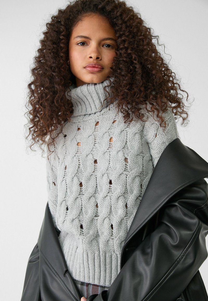 Giovane donna con capelli ricci che indossa un maglione a collo alto grigio chiaro a trecce e una giacca di pelle nera appoggiata sulle spalle.