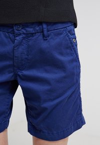 Detailní záběr na osobu, která má na sobě jednobarevné modré volné šortky s knoflíkem, zipem, poutky na opasek a bočními kapsami, v kombinaci s černým topem.