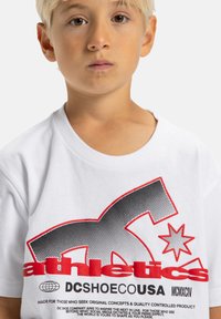 Biele bavlnené tričko s čiernym a červeným grafickým textom a dizajnom. Text obsahuje slová "athletics" a "DCSHOECOUSA." Poskytuje okrúhly výstrih a krátke rukávy.