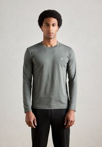 T-shirt a maniche lunghe verde oliva in tessuto morbido con scollatura rotonda e piccolo logo bianco sul lato sinistro del petto. Indossato con pantaloni neri.