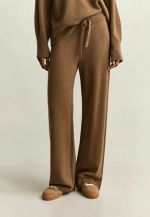 Personne portant un pantalon en maille marron ample avec une taille à cordon, un pull marron assorti et des chaussures marron à enfiler, debout sur un fond neutre.