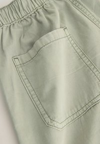 Shorts en tissu vert clair avec une ceinture élastique, dotés d'une poche arrière unique et de détails de couture visibles. Surface douce et texturée.