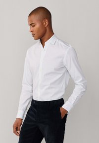 Witte blouse met een subtiel stippenpatroon, lange mouwen en een nette kraag, gecombineerd met donkere, tonale broeken. Glad stoftextuur.