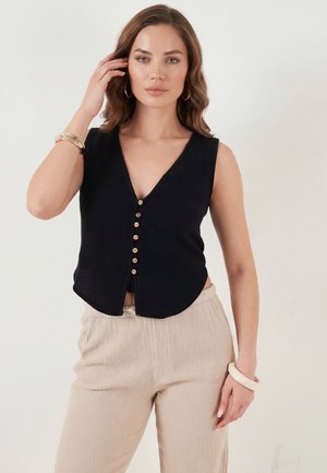 Kvinde med skulderlangt brunt hår iført sort ærmeløs knapbluse og beige teksturerede bukser, poserende med hænden nær ansigtet.