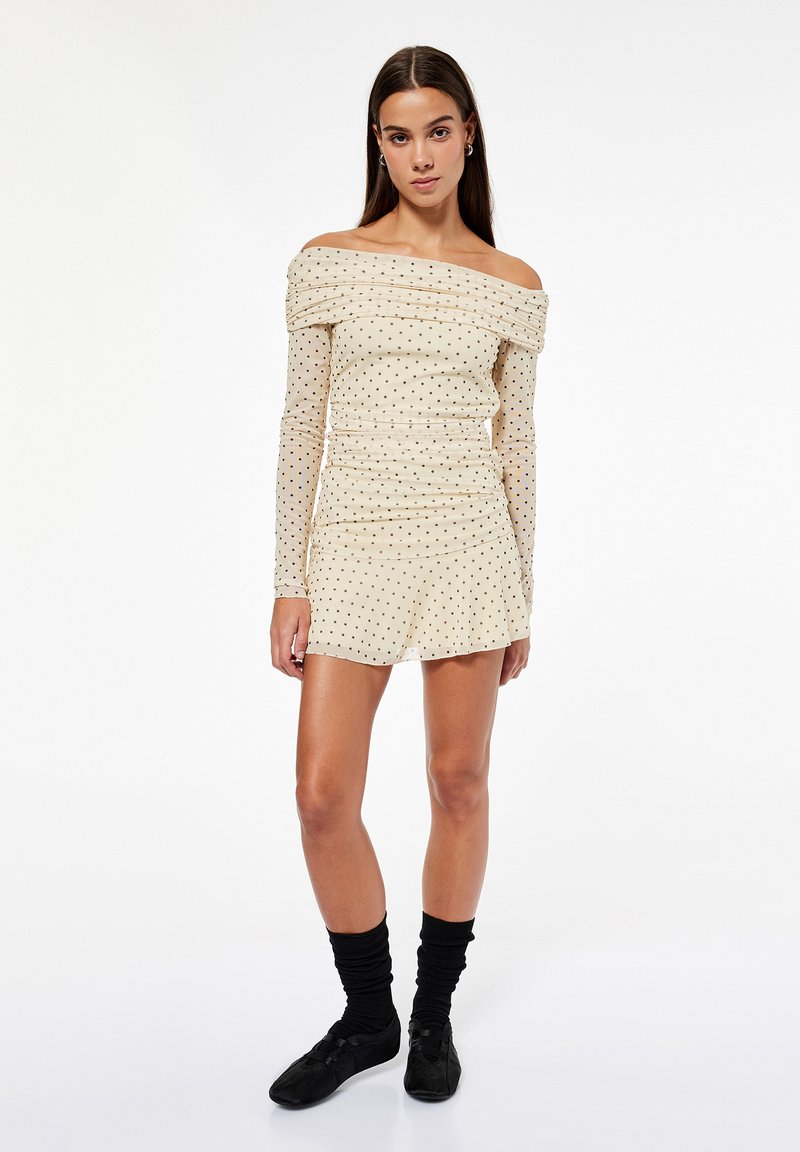 Donna che indossa un vestito crema con spalle scoperte, pois neri e maniche lunghe, abbinato a calzini neri e scarpe stile balletto nere.