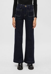 Sötétkék, bő szárú farmernadrág, amely denimből készült. Két elülső zsebbel, kontrasztos sárga varrással és magas derékra, fém gombos záródással rendelkezik.