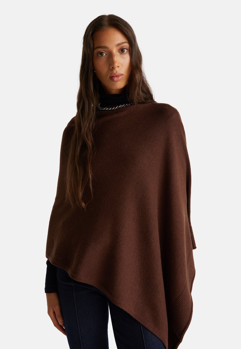 Hnědý pletený pončo s jemnou texturou, s asymetrickým lemem a širokým střihem, vrstvené přes tmavý rolák.