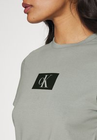 Camiseta de algodón gris con un logotipo rectangular negro con letras blancas "cK". Cuello redondo y mangas cortas. Textura suave, forma ajustada.