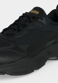 Sort Puma-sneaker med tekstureret og glat syntetisk materiale, sorte snørebånd og gyldent Puma-logo på tungen.