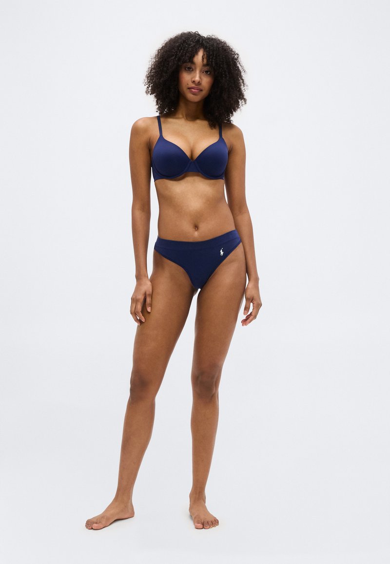 Femme aux cheveux bouclés portant un soutien-gorge bleu marine et un sous-vêtement bikini assorti avec un petit logo blanc, debout pieds nus sur fond blanc.