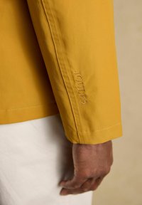 Rechterarm met een mosterdgele jas met "JOULES" geborduurd vlak bij de pols, tegen een beige achtergrond.