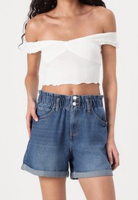 Ung kvinna klädd i vit croptop med avskuren axel och högmidjade uppvikta denimshorts med elastiskt midjeband och framknappar.