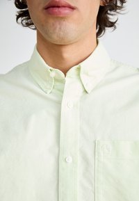 Lys grøn skjorte med knapper laget av bomull, med krage, frontlomme og hvite knapper. Stoffet har en litt teksturert overflate.