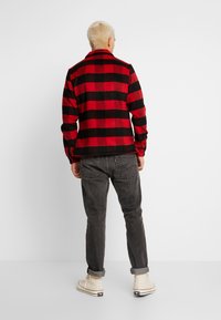 Camicia in flanella a scacchi rossa e nera con colletto, maniche lunghe e texture morbida, abbinata a jeans scuri e sneakers beige alte.