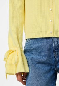 Gul ribbad kofta med pärlknappar; har vida ärmar som knyts vid handlederna. Bärs över blå jeans.