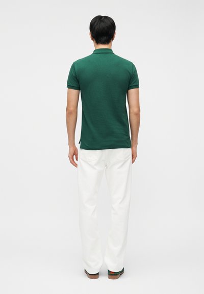 Άνδρας που στέκεται με την πλάτη στην κάμερα, φορώντας σκούρο πράσινο πόλο με κοντά μανίκια και άσπρο παντελόνι με ίσια μπατζάκια, μπροστά σε απλό φόντο.