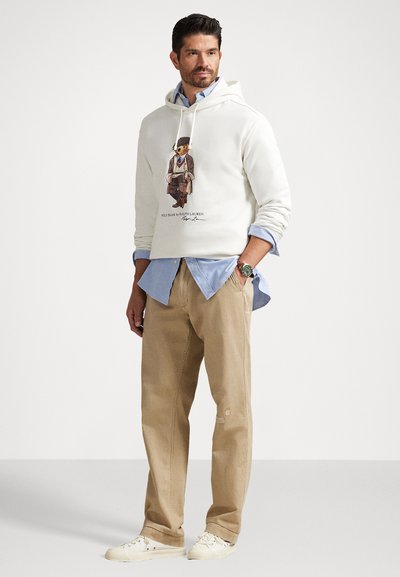 Homem a usar calças beije, ténis brancos, camisa azul clara sob um moletom branco com um gráfico de um urso, de pé com uma mão no bolso contra um fundo branco.